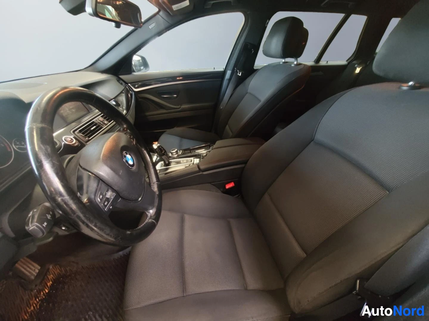 Muu: Auton sisätilat kuljettajan puolelta: BMW-ohjauspyörä, tumma kangasverhoilu, etuistuimet ja keskikonsoli näkyvät.