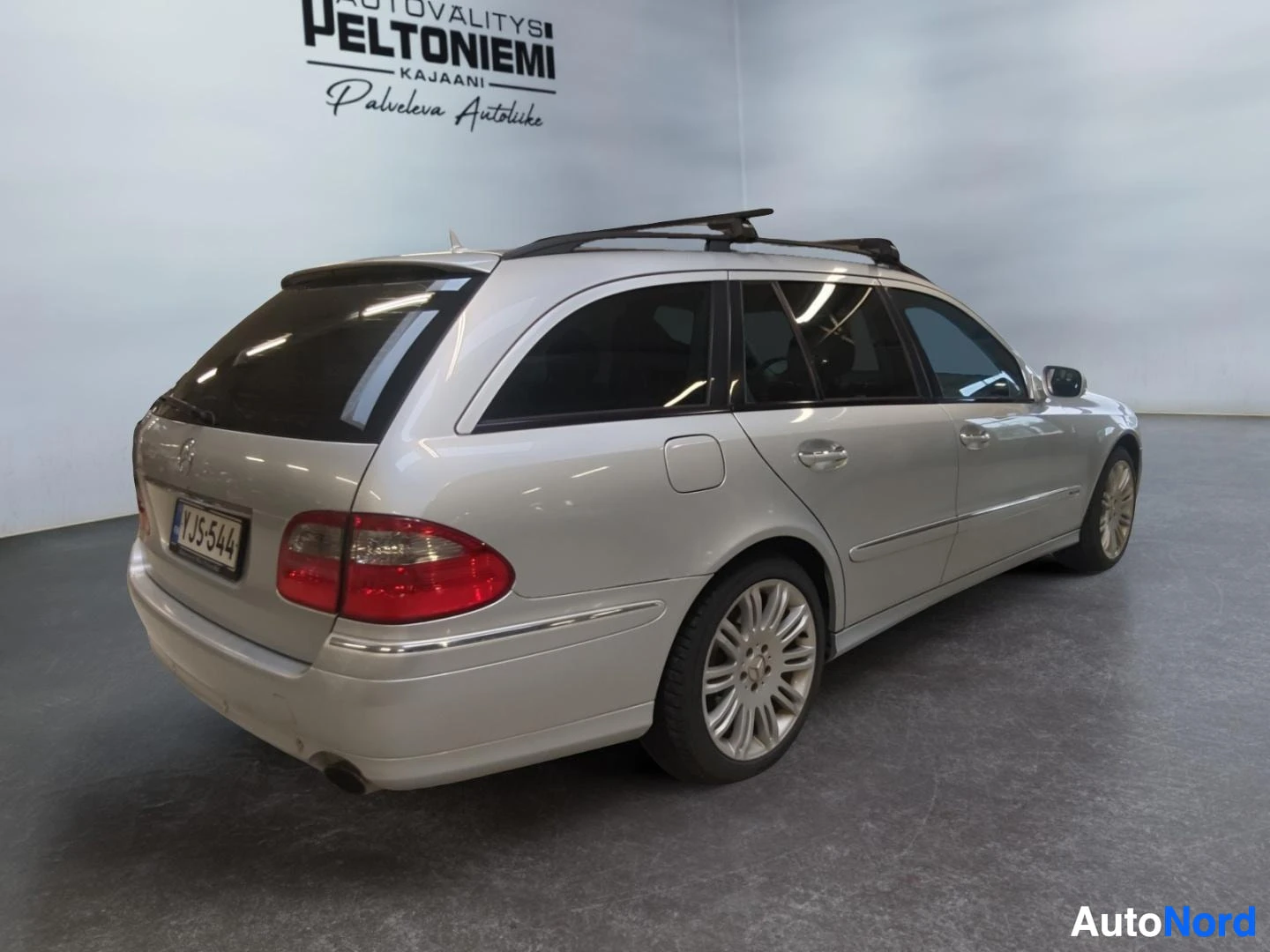 Takaviisto: Farmarityylinen auton takaosa ja sivu kulmasta kuvattuna; hopeakorinen_auto tummissa ikkunoissa ja kattotelineet sisätiloissa.