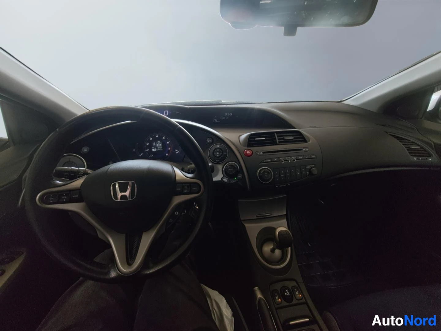 Edestä: Näkymä ohjaamosta tuulilasin suuntaan: Honda-merkitty ohjauspyörä, mittaristo ja keskusyksikkö. Kuva on peiliin heijastunut, oikea reuna heijastaa sisäpeilin re