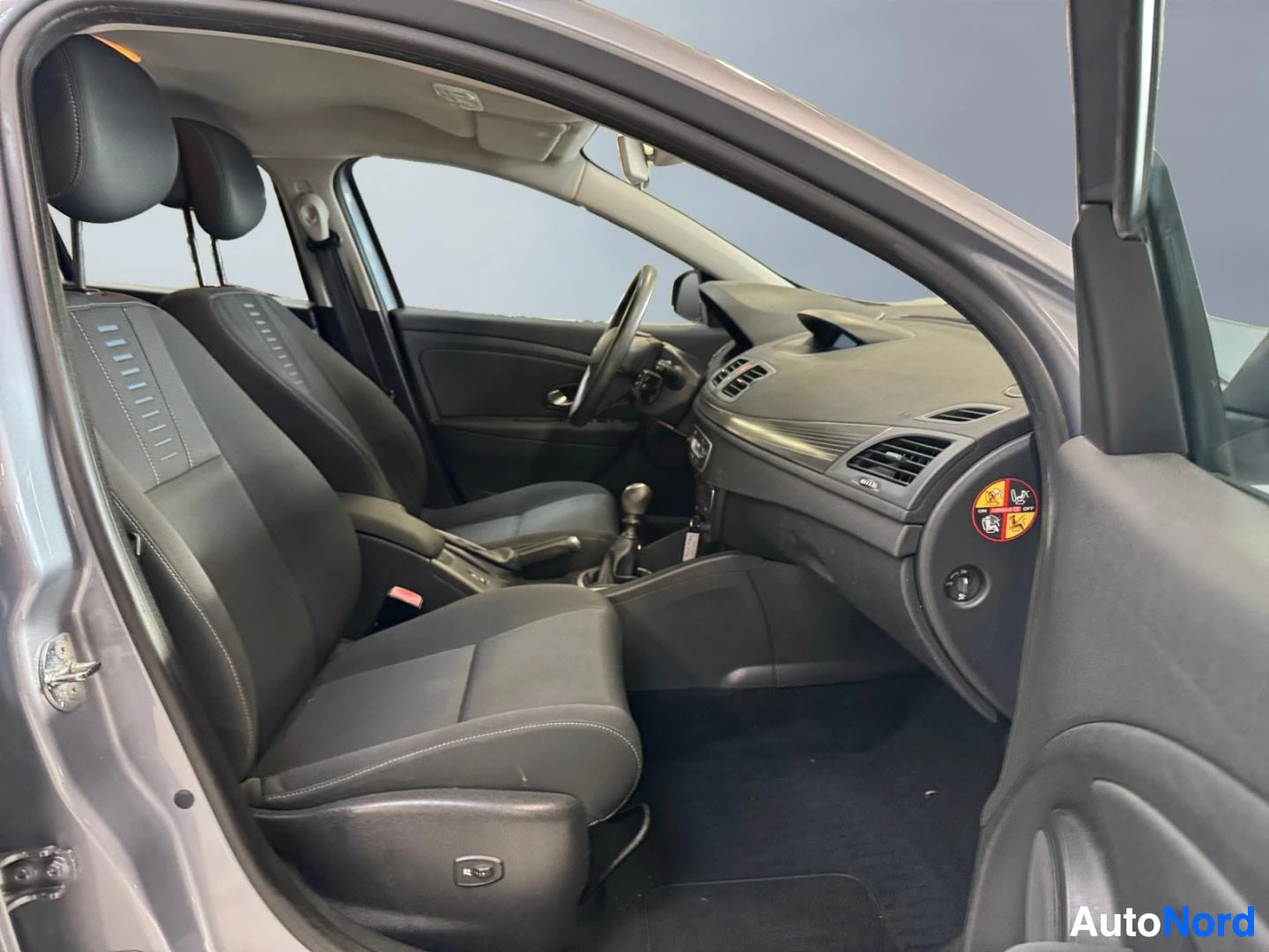 Muu: Renault Meganein etumatkustustila kuvattuna kuljettajan oven aukosta: tummat kangasistuimet, ohjauspyörä ja manuaalivaihtopeksi.