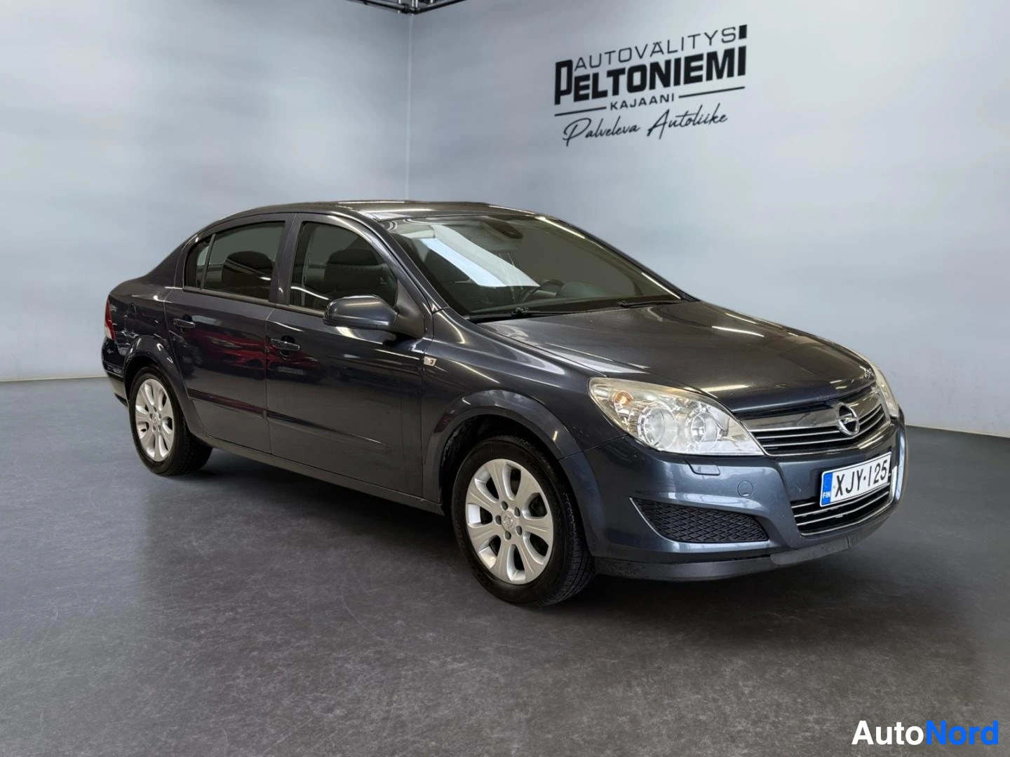 Etuviisto, nokka oikealle: Opel Astra -henkilöauto tummansinisenharmaana etukulmakuva sisätiloissa, rekisterikilpi.