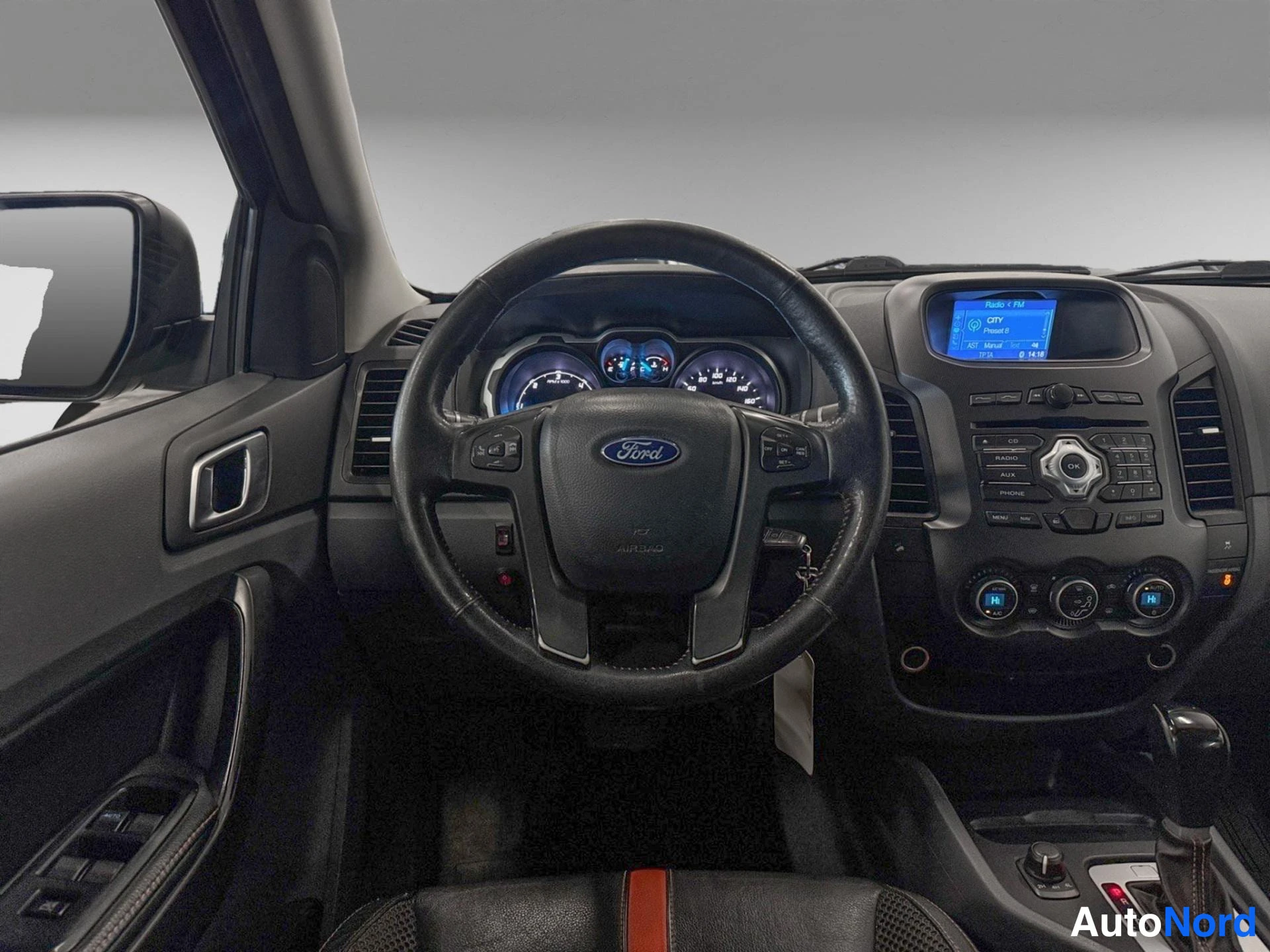 Edestä: Auton ohjaamo: Ford-merkkinen ohjauspyörä, mittaristo, keskikonsolin radio-näyttö ja automaattivaihteen valitsin.