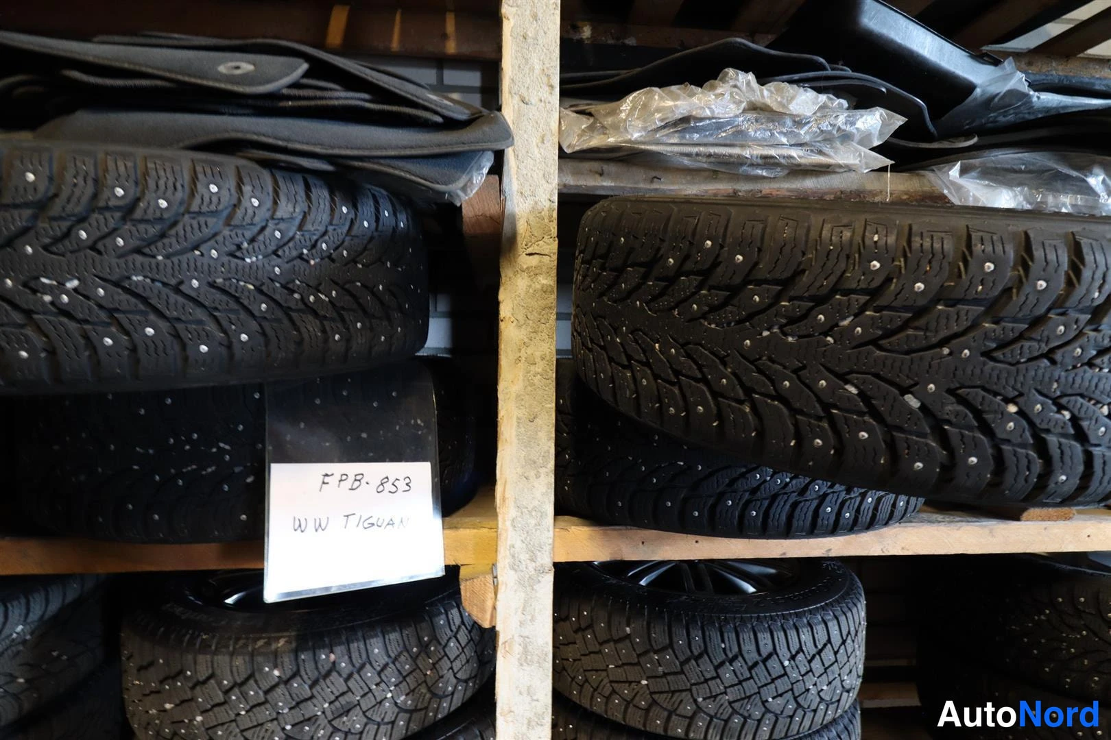 Muu: Hyllyllä pinottuna nastarenkaita, joukossa käsin kirjoitettu lappu tekstillä WW TIGUAN.