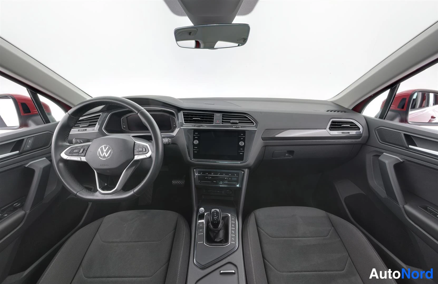 Yksityiskohta: Volkswagenin ohjaamo suoraan edestä: Volkswagen-merkitty ohjauspyörä, kojelauta, kosketusnäyttö ja automaattivaihteiston valitsin, tummat istuimet.