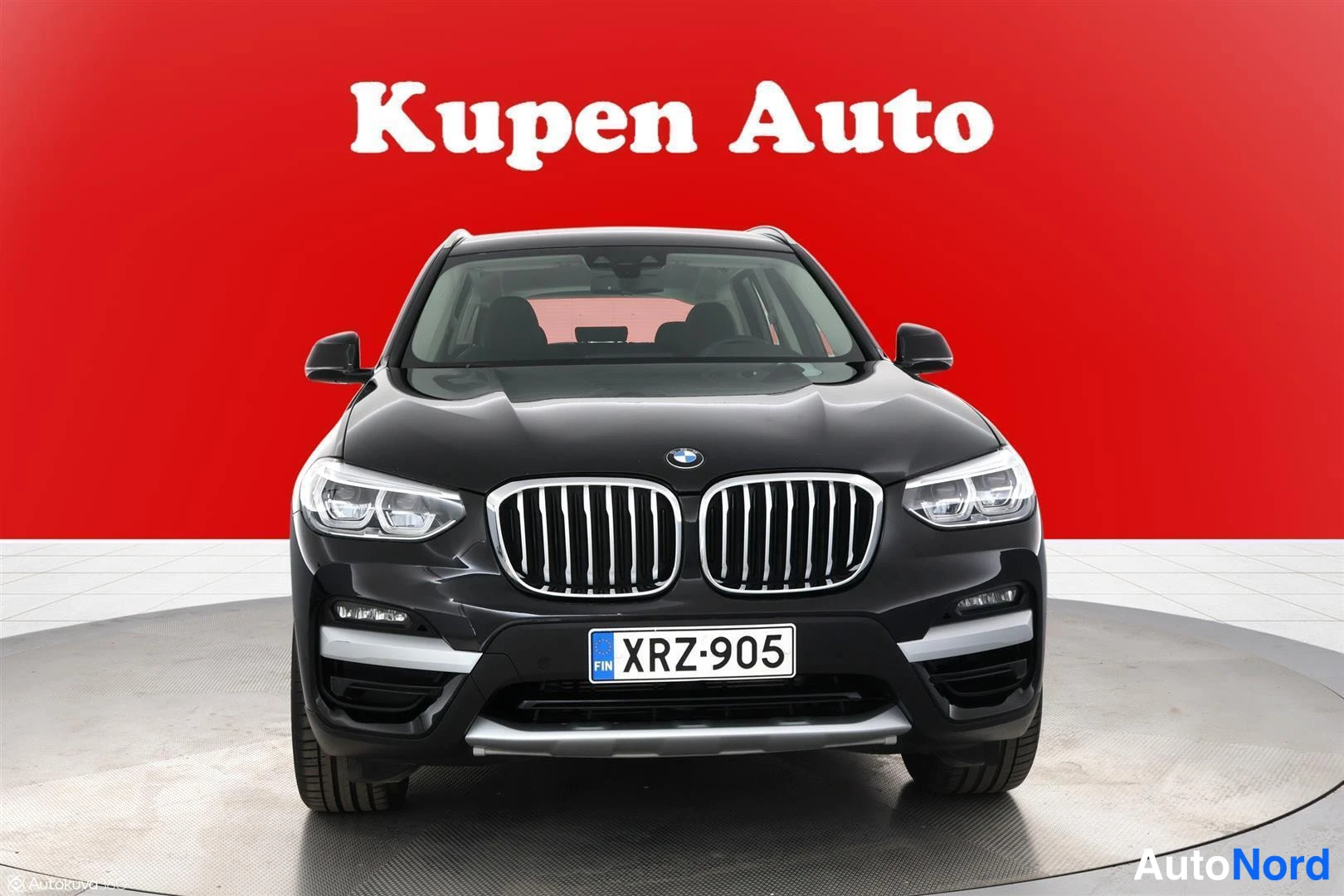 Edestä: Musta BMW X3 suoraan edestä valokuvattu, rekisteritunnus, punainen tausta.