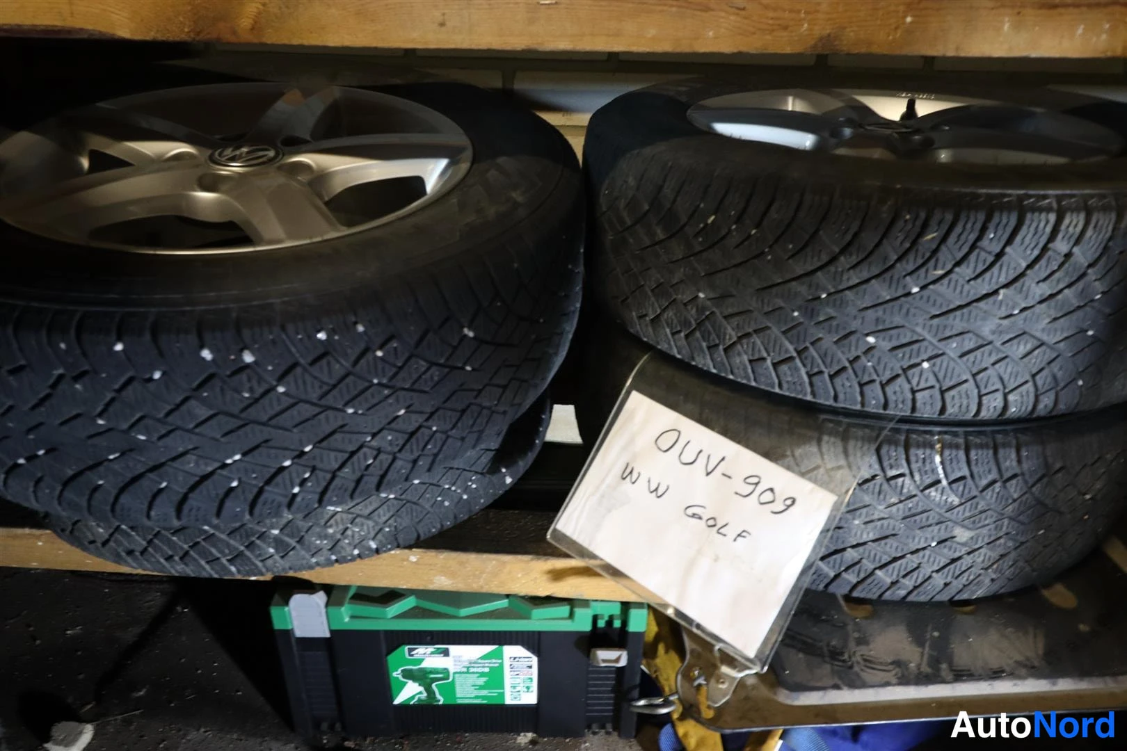 Muu: Vararenkaat tai talvirenkaat hyllyllä: kaksi nastarengasta vanteilla ja käsinkirjoitettu lappu ' VW GOLF'.