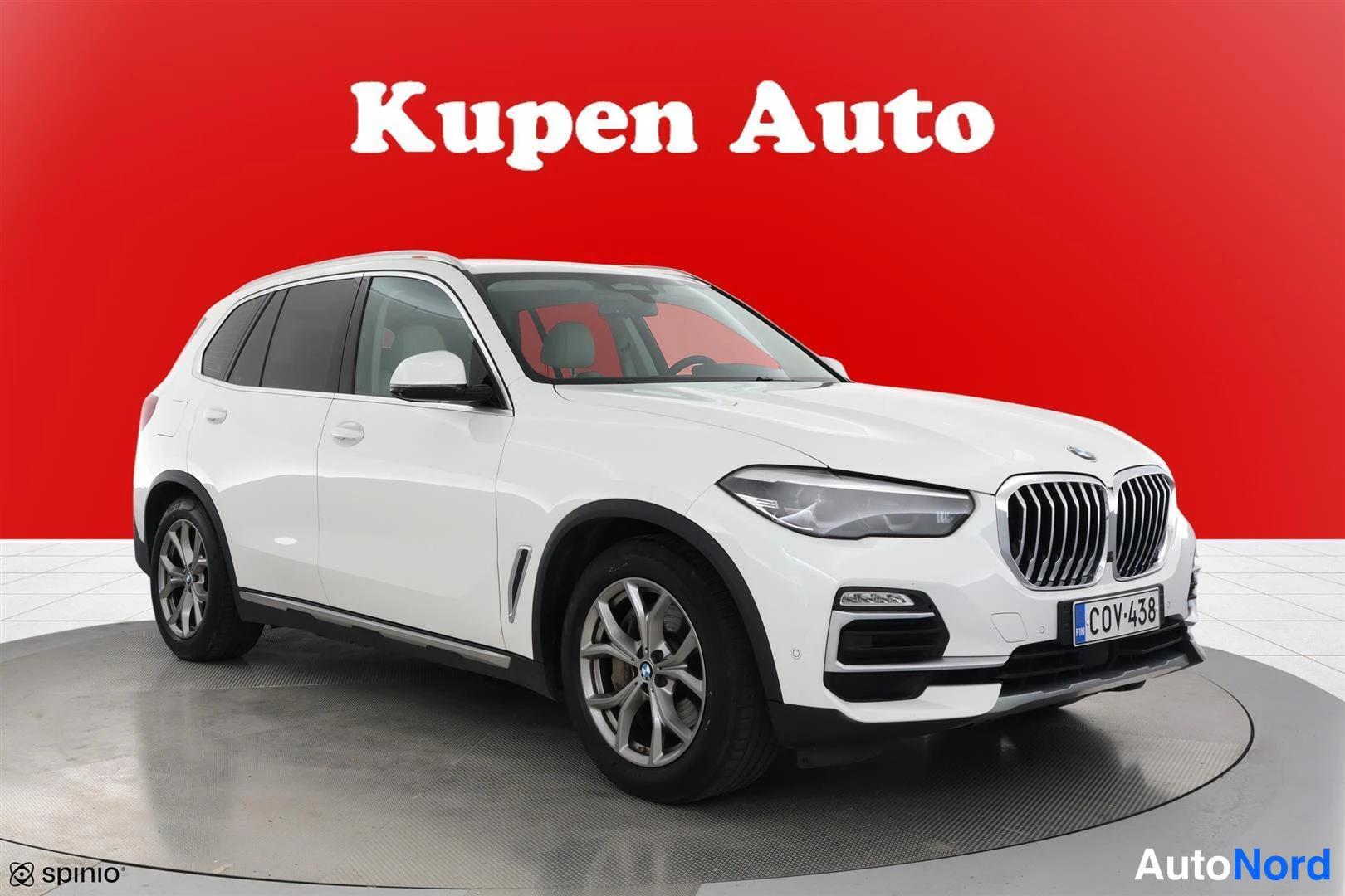 Etuviisto, nokka oikealle: Valkoinen BMW X5 neljästä etukulmasta punaisella taustalla, Kupen Auto -mainosteksti ylhäällä.