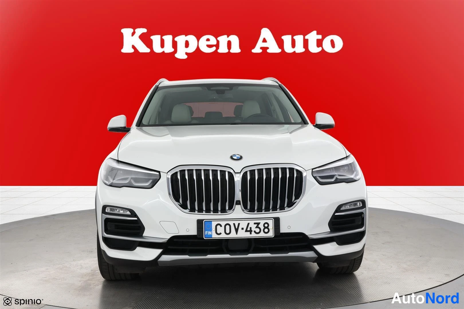 Edestä: Valkoinen BMW X5 suoraan edestä, BMW-merkki ja etusäleikkö keskitettynä punaisella taustalla.