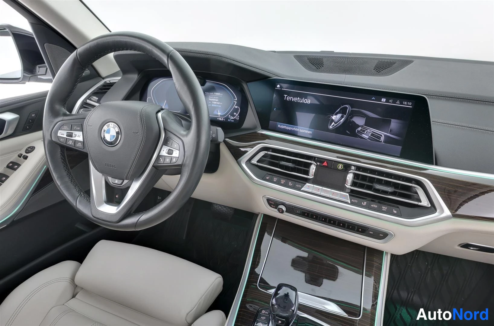 Edestä: Kuljettajan puolen sisätila: BWM-ohjauspyörä, elektroninen kojelauta, suuri keskinäyttö tekstillä 'Tervetuloa' ja vaaleat penkit.