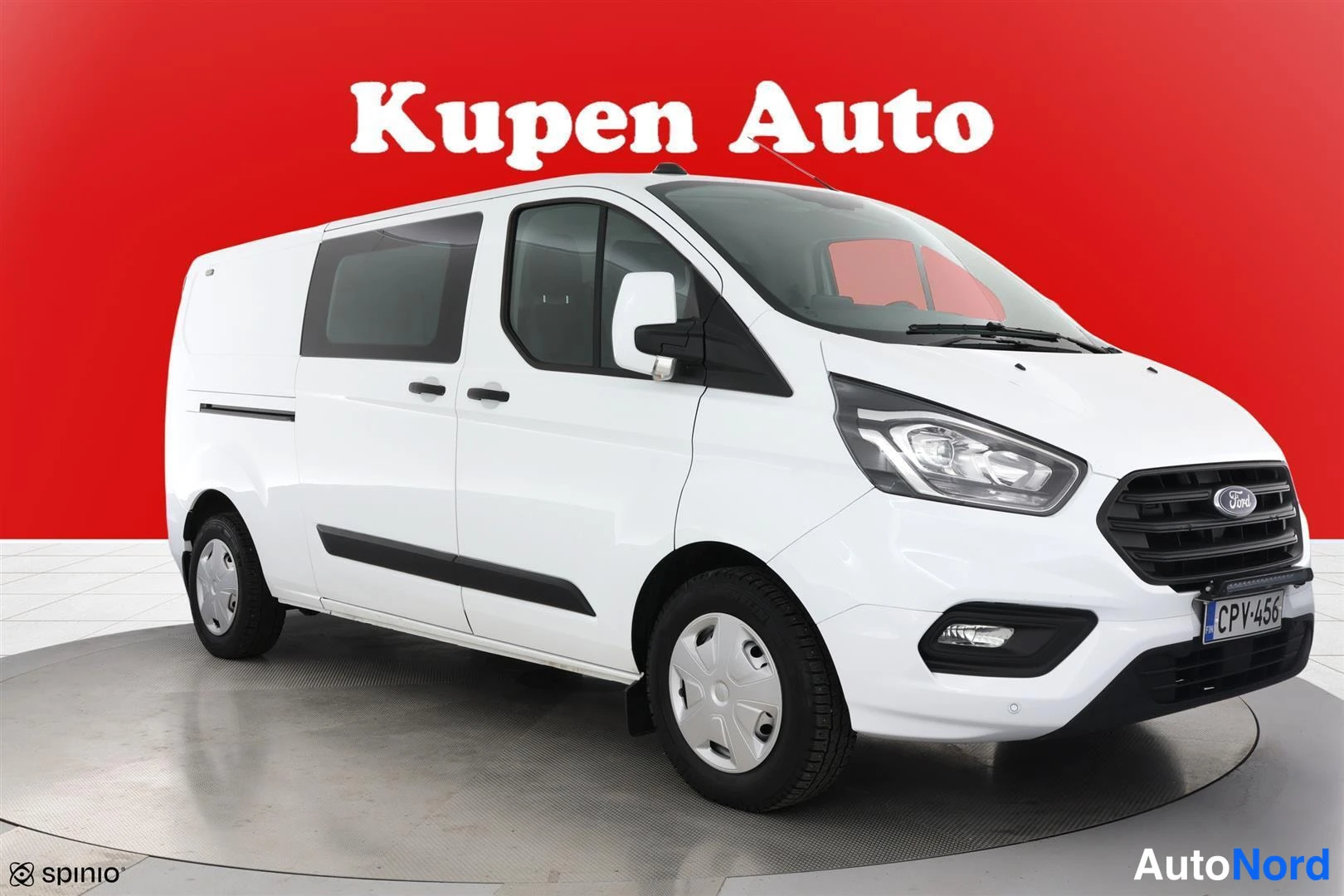 Etuviisto, nokka oikealle: Valkoinen Ford Transit Custom -pakettiauto kulmasta oikealta etupää vasemmalla punaisella taustalla