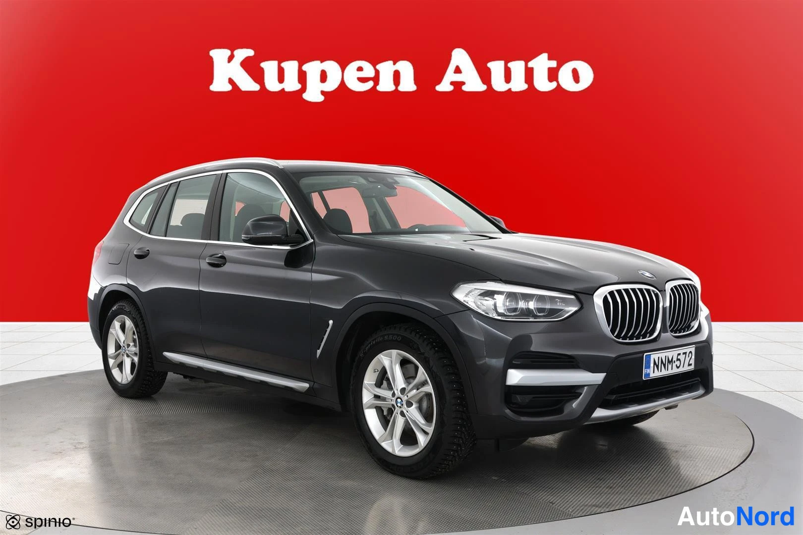 Etuviisto, nokka oikealle: Tummanharmaa BMW X3 farmari punaisella taustalla, kolme neljäsosaa edestä oikealta.