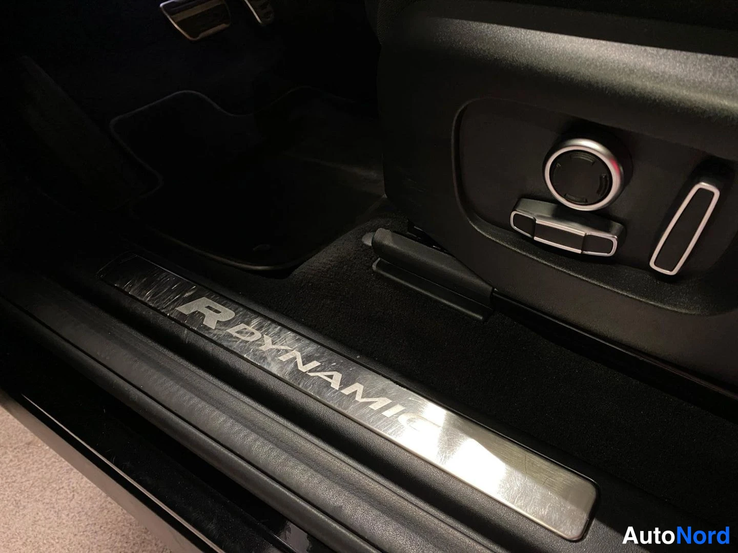 Yksityiskohta: Kynnyslistassa R-DYNAMIC-teksti, istuinten säätönapit ja matto oikealla etuistuimen alla.