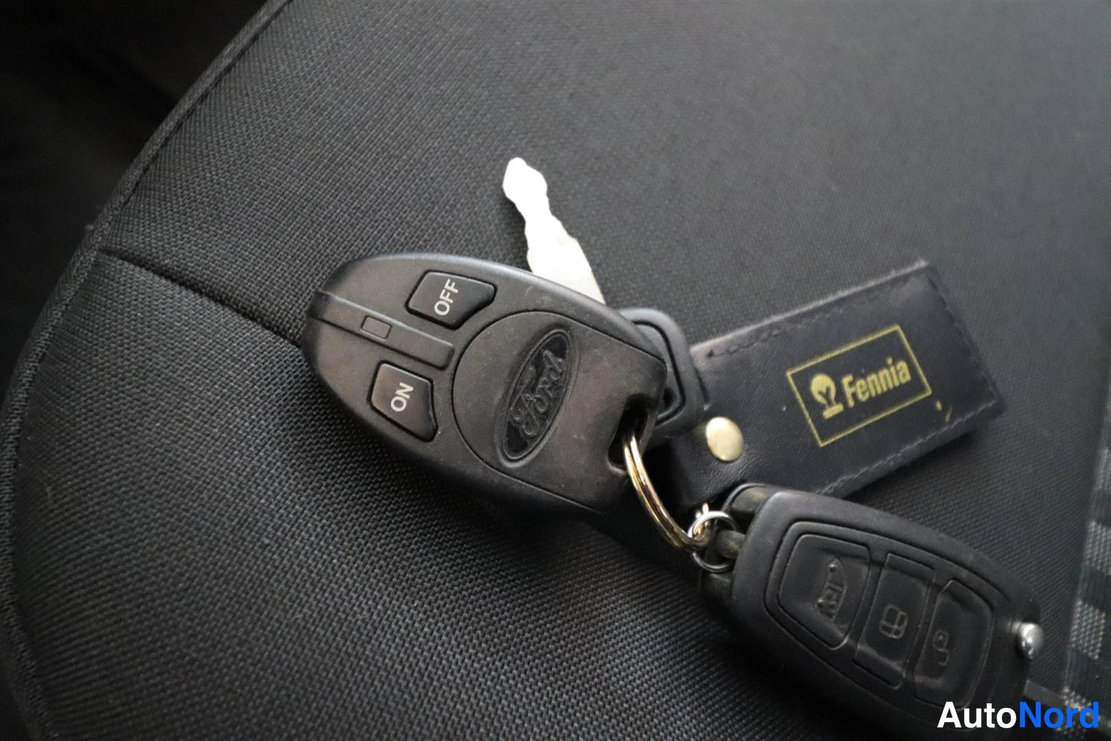 Yksityiskohta: Kaksi Ford-avainparia, joissa etäohjaimet, sekä Fennia-avainnappi tummalla penkillä.