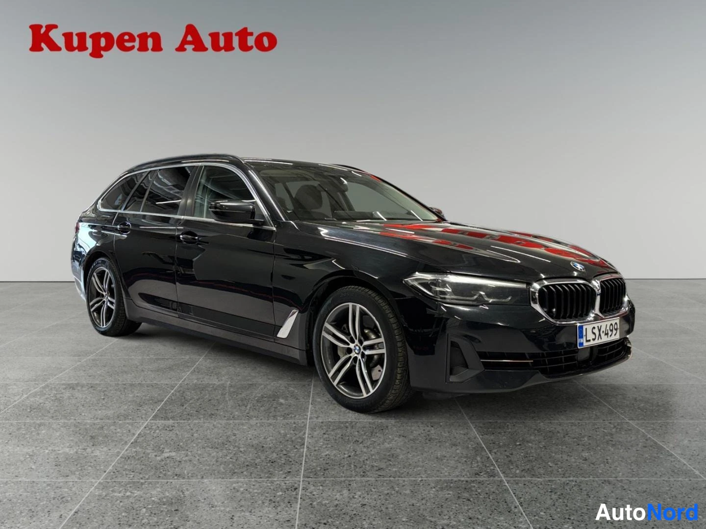 Etuviisto, nokka oikealle: Musta BMW -farmari etukulmasta oikealta, rekisteritunnus, Kupen Auto -logo kuvassa.