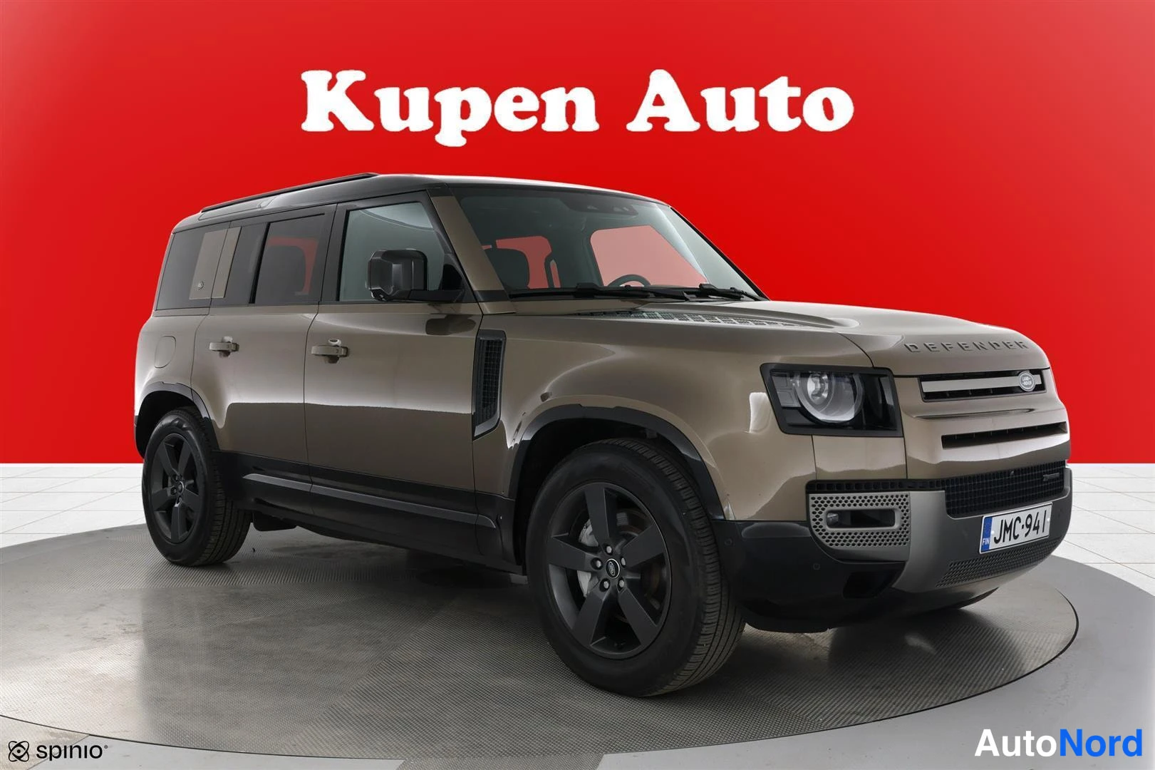 Etuviisto, nokka oikealle: Land Rover Defender etuviisto oikealta, ruskeansävyinen korimalli tummilla vanteilla punaisella taustalla.
