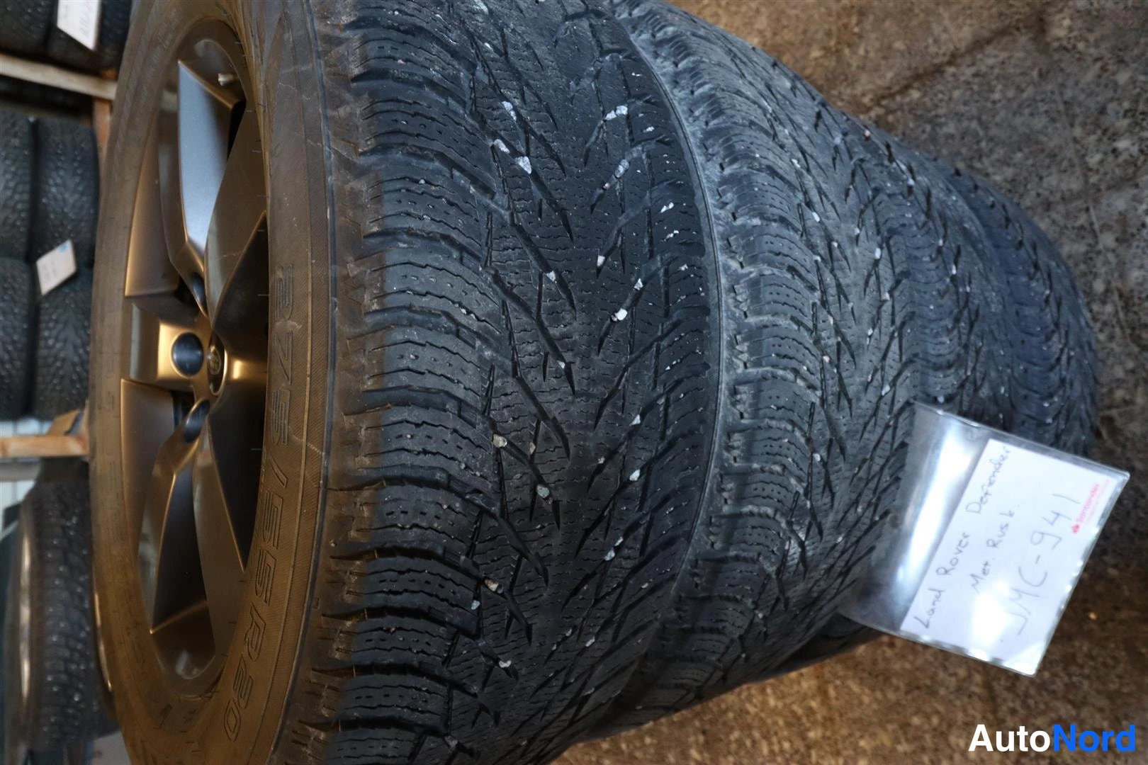 Yksityiskohta: Talvirenkaita vanteilla pinossa, mukana käsin kirjoitettu laput, jossa lukee Land Rover Defender ja rekisteritunnus.