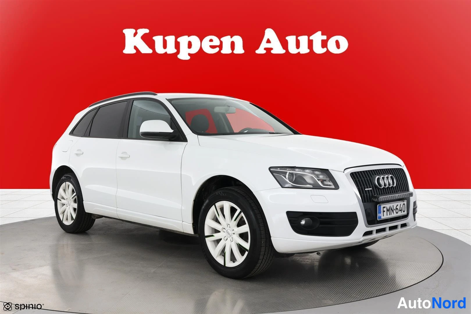 Etuviisto, nokka oikealle: Valkoinen Audi Q5 -henkinen farmarityyppinen henkilöauto kuvattuna etukulmasta, keula oikealla, rekisterikilpi, punainen tausta.