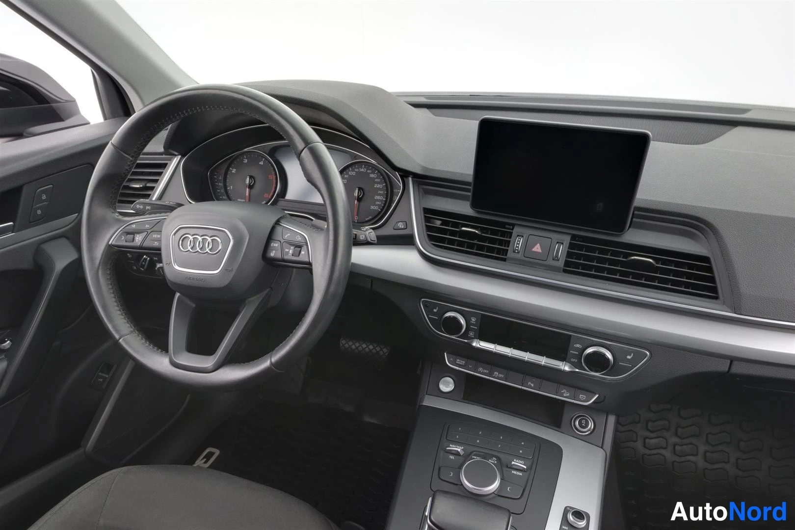 Ohjaamo: Ohjauspyörän ja kojelaudan lähikuva; Audi-merkki, mittaristo ja keskinäyttö näkyvissä.
