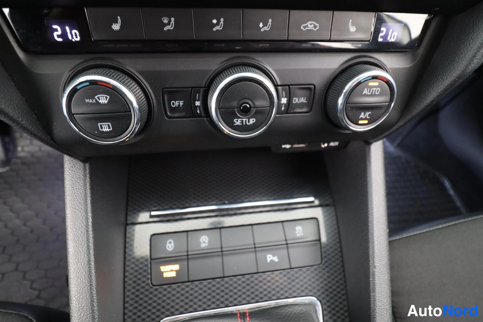 Yksityiskohta: Auton kojelaudan ilmastointipaneeli: kolme säätöpyörää, DUAL- ja AUTO/AC-merkit, lämpötilat 21,0 molemmin puolin sekä alla pysäköintiapikytkimet.