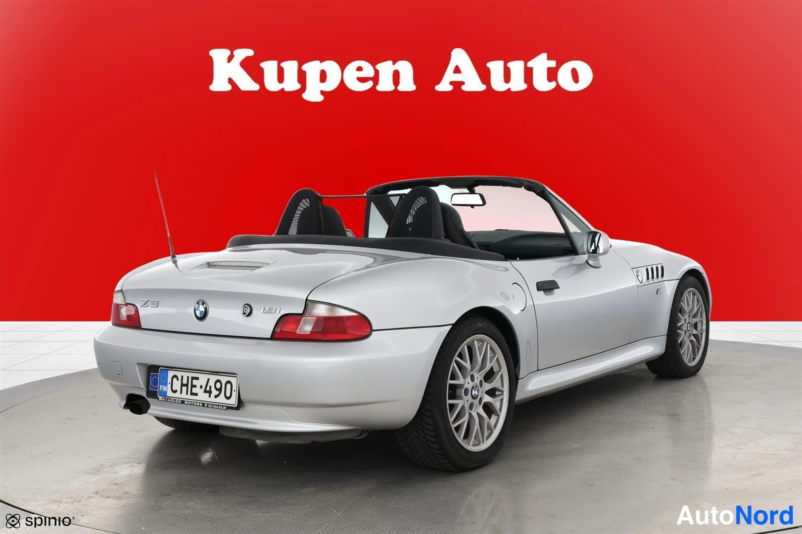 Takaviisto: Hopeanvärinen avokattoinen BMW Z3 -urheiluauto takaa sivusta kuvattuna punaisella taustalla.