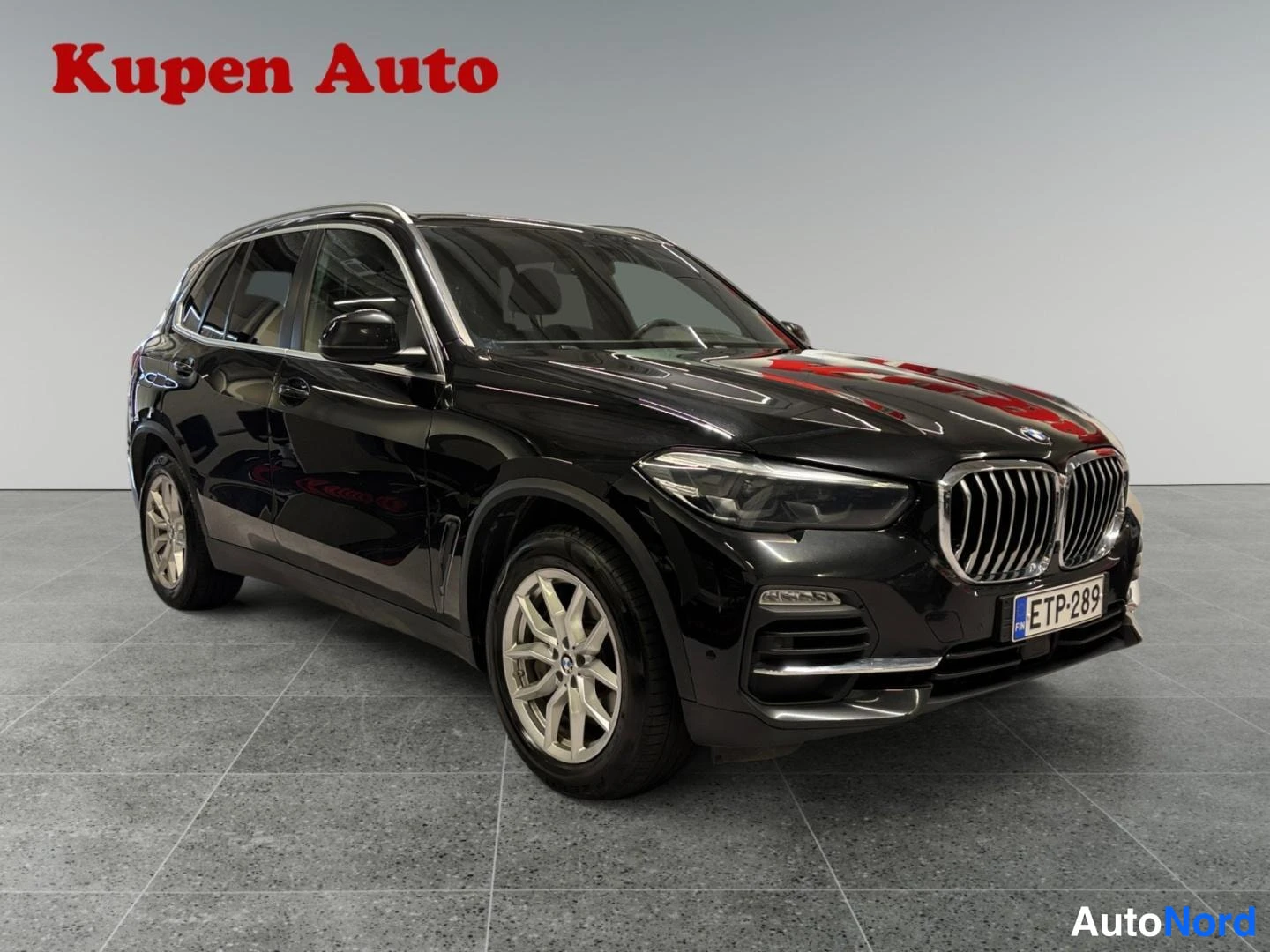 Etuviisto, nokka oikealle: Musta BMW X5 -maastauto kulmasta edestä valokuvattuna sisätiloissa, vanteet ja rekisterikilpi näkyvissä.