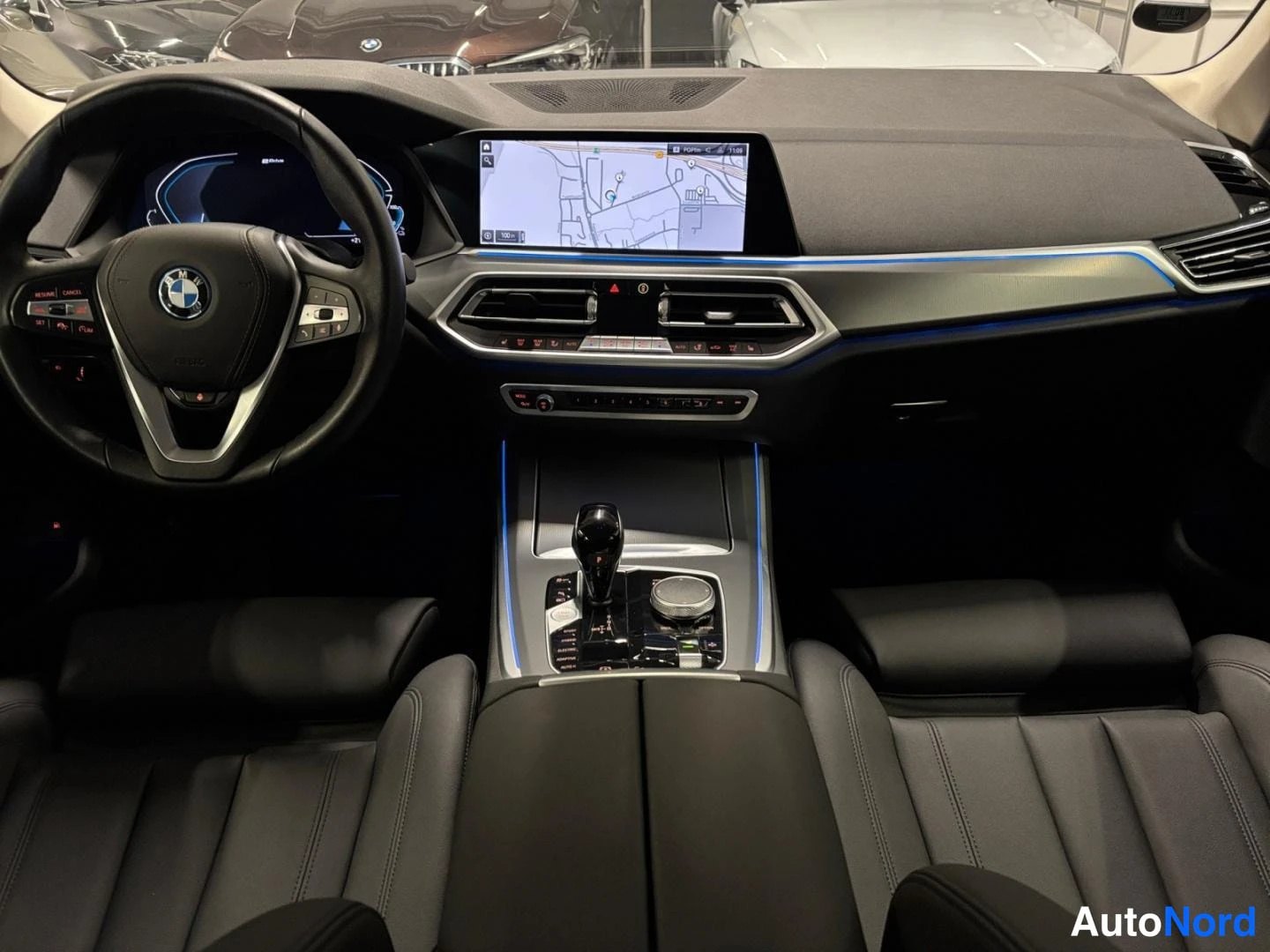 Muu: Ohjaamo nähtynä keskeltä: BMW:n ohjauspyörä, digitaalinen mittaristo, kartta kesk näytöllä ja automaattivaihteen valitsin keskikonsolissa.