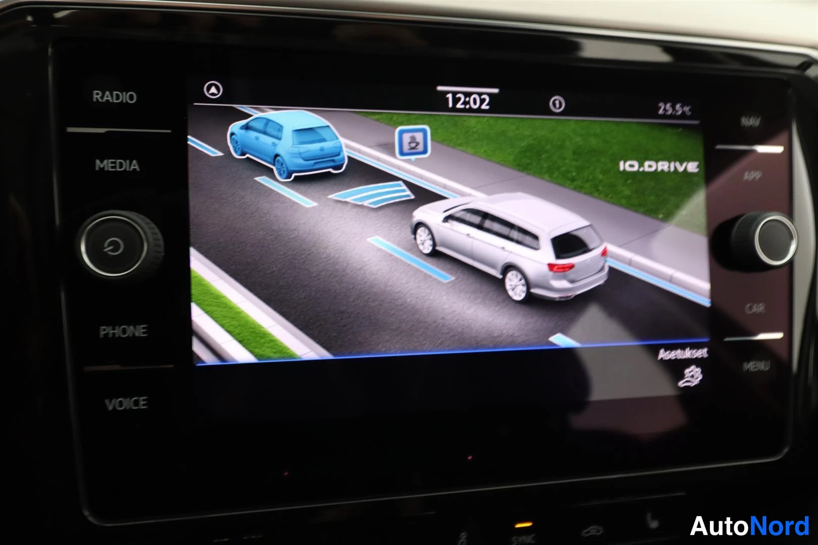 Muu: Infotainment-näyttö, jolla näkyy kuvituskuva autosta ja toisesta ajoneuvosta, kellonaika 12:02.