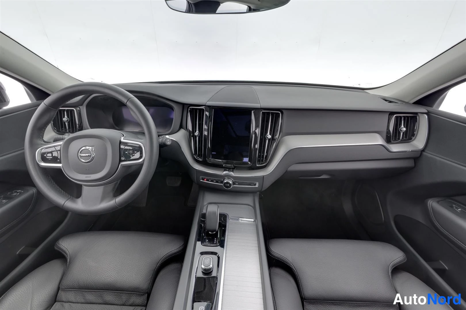 Muu: Auton ohjaamo edestä kuvattuna: Volvo-ohjauspyörä, keskikonsolin kosketusnäyttö, automaattivaihteen valitsin ja kaksi tummaa etuistuinta.