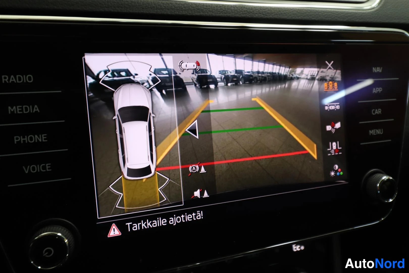 Yksityiskohta: Infotainment-näytöllä pysäköintikameran kuva ja ympäriajonäkymä, varoitus tekstillä 'Tarkkaile ajetietä!'.