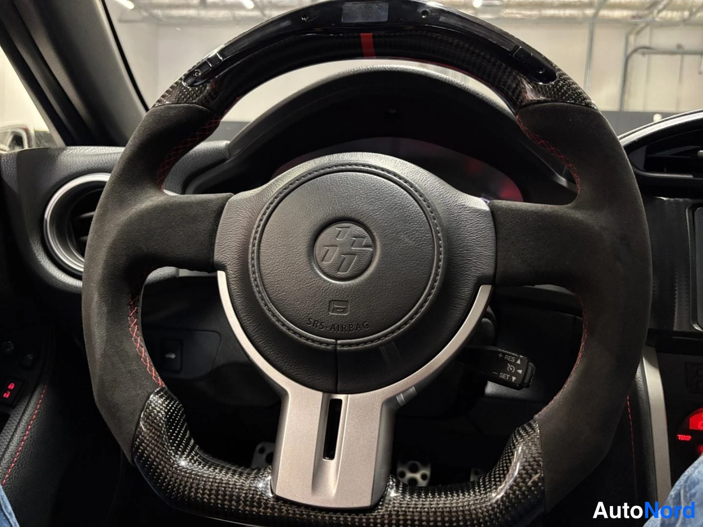 Muu: Ohjauspyörä lähikuvassa: hiilikuitureunus, mokkanahkapinta punaisilla ompeleilla, SRS AIRBag -teksti ja Toyota 86 -merkki nappikeskuksessa.