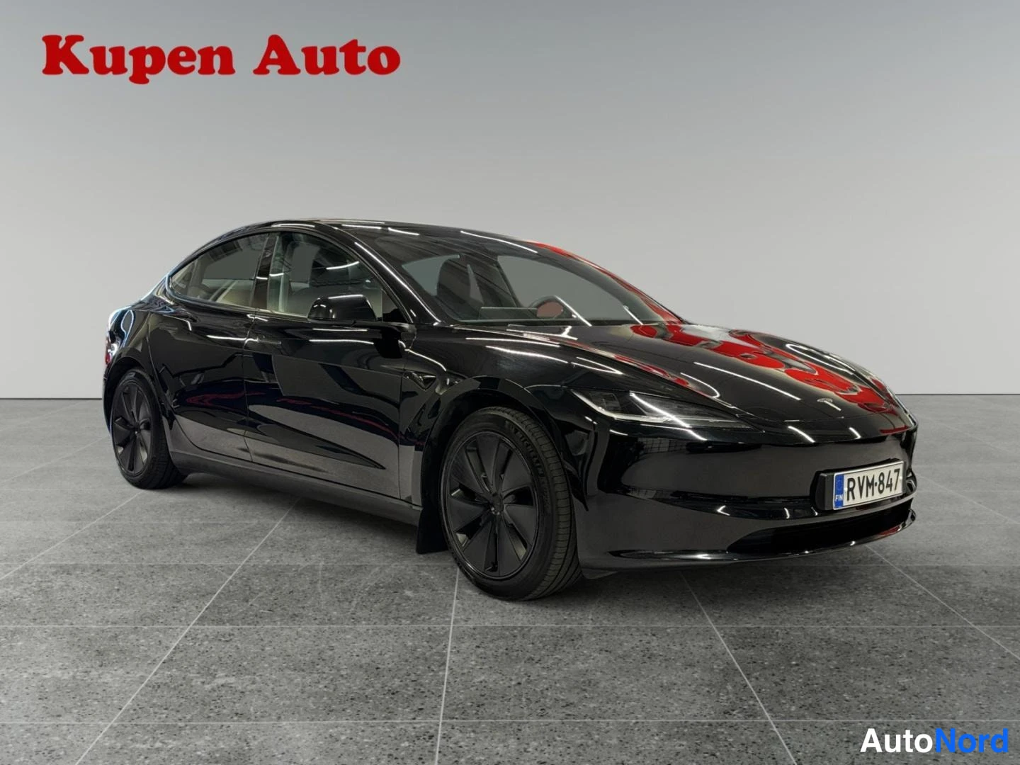 Etuviisto, nokka oikealle: Musta Tesla Model 3 -henkilöauto etukolmannesviistosta vasemmalta sisätiloissa, rekisteritunnus.