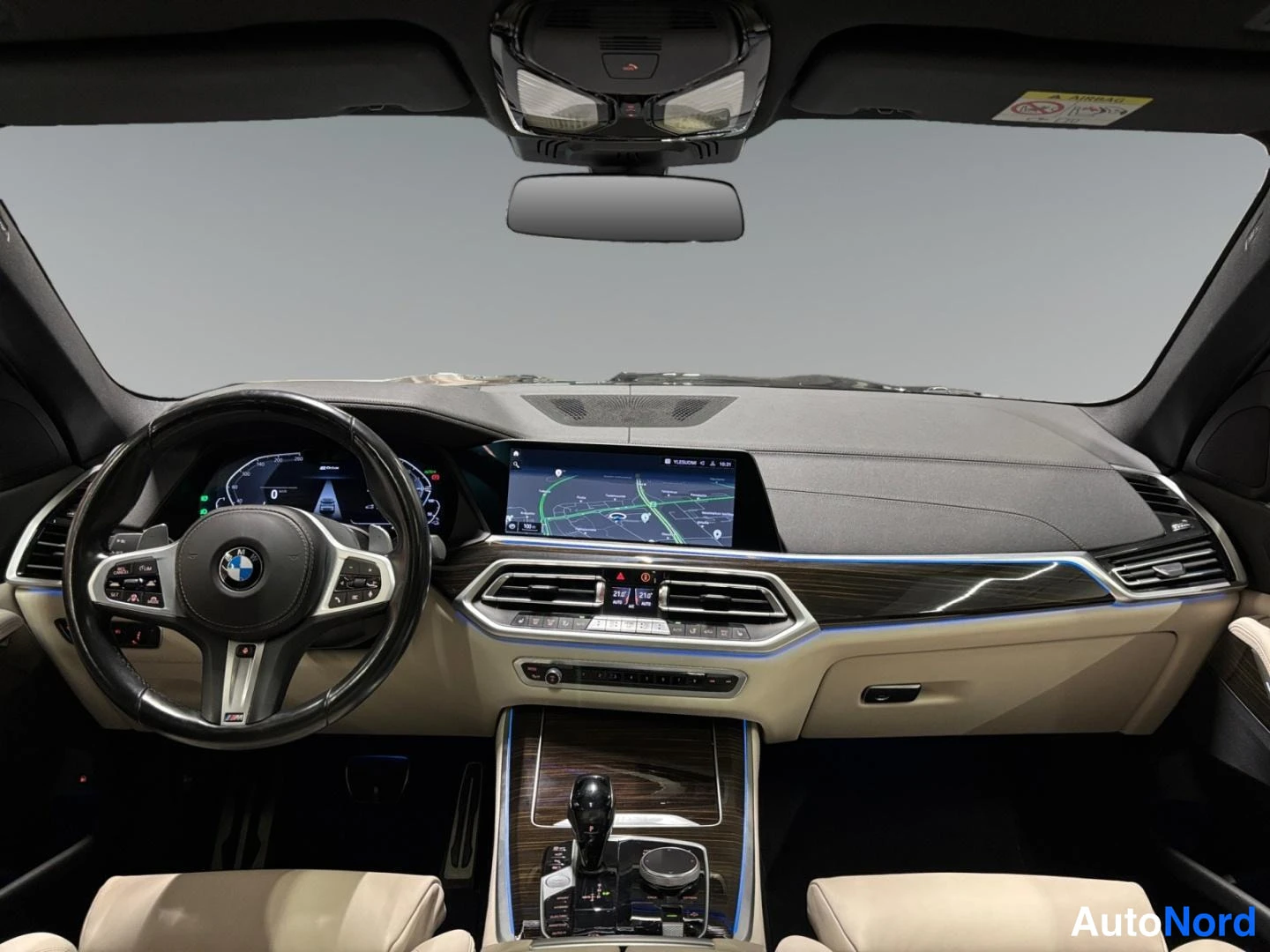 Muu: Sisätilakuva: BMW:n ohjaamo beigellä nahkaistuimilla, tumma kojelauta, infotainment-näyttö kartalla, M-ohjauspyörä ja puukuvioidut koristeosat.