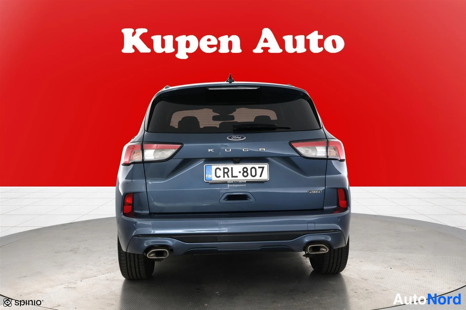Takaa: Sinisenharmaan tila-auton takaosa suoraan takaa: KUGA-kirjaimet, rekisterikilpi, kaksi pakoputkea ja punainen Kupen Auto -tausta.
