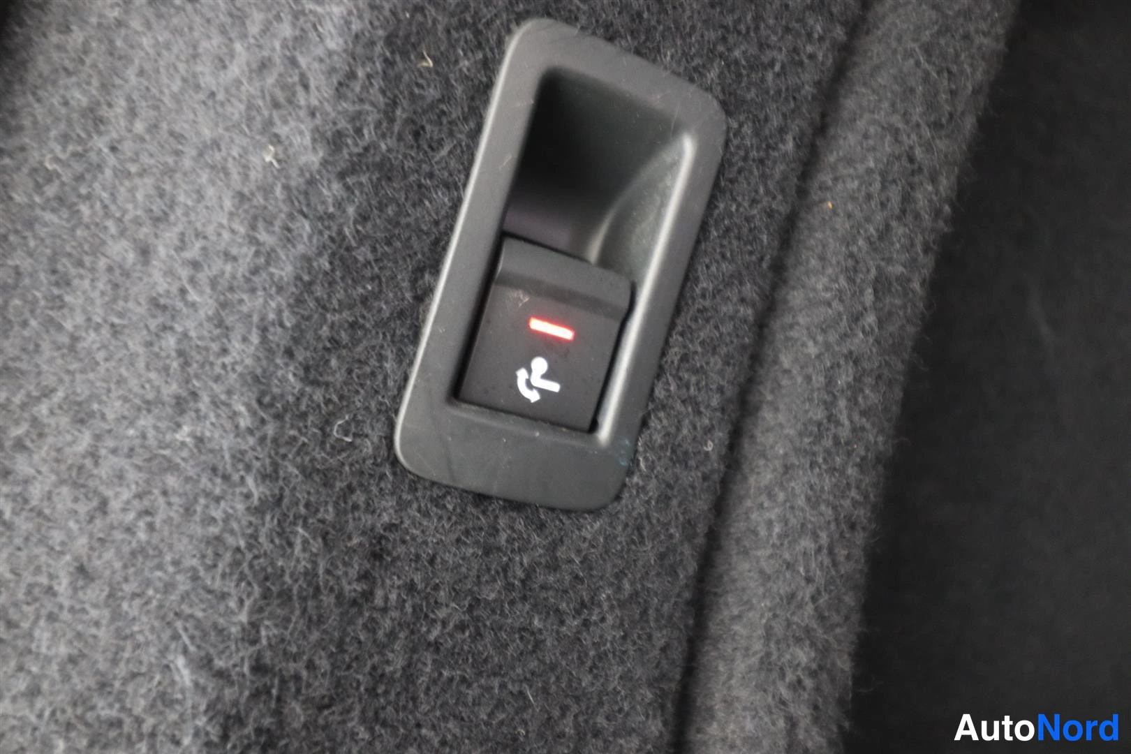 Yksityiskohta: Tumma painike punaisella merkkivalolla ja istuinta esittävällä symbolilla pehmeässä harmaassa kankaassa.