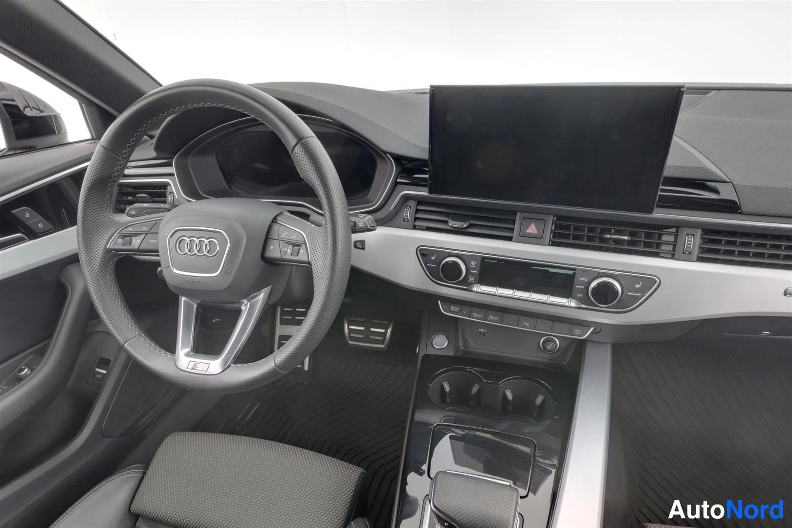 Muu: Kuljettajan puolen ohjaamo lähikuvassa: Audi-ohjauspyörä, iso keskinäyttö, ilmastointilaitteiden pyörät ja vaihteenvalitsin näkyvät.