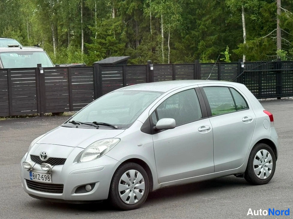 Etuviisto, nokka vasemmalle: Hopeanvärinen Toyota Yaris viistosti edestä vasemmalta aidan vieressä; konepellillä kaksi lisävaloa, rekisteri.