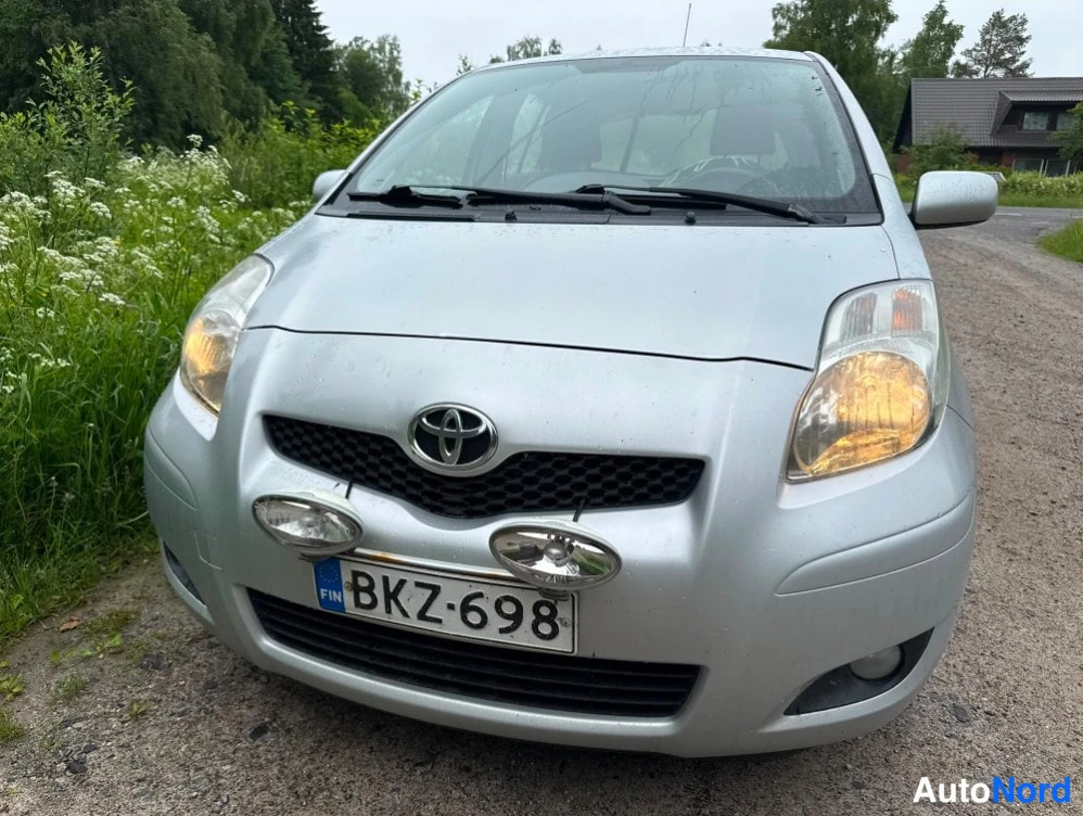 Edestä: Etuperspektiivi hopeanvärisestä Toyota Yariksesta; konepellillä kaksi lisävaloa, rekisteri.