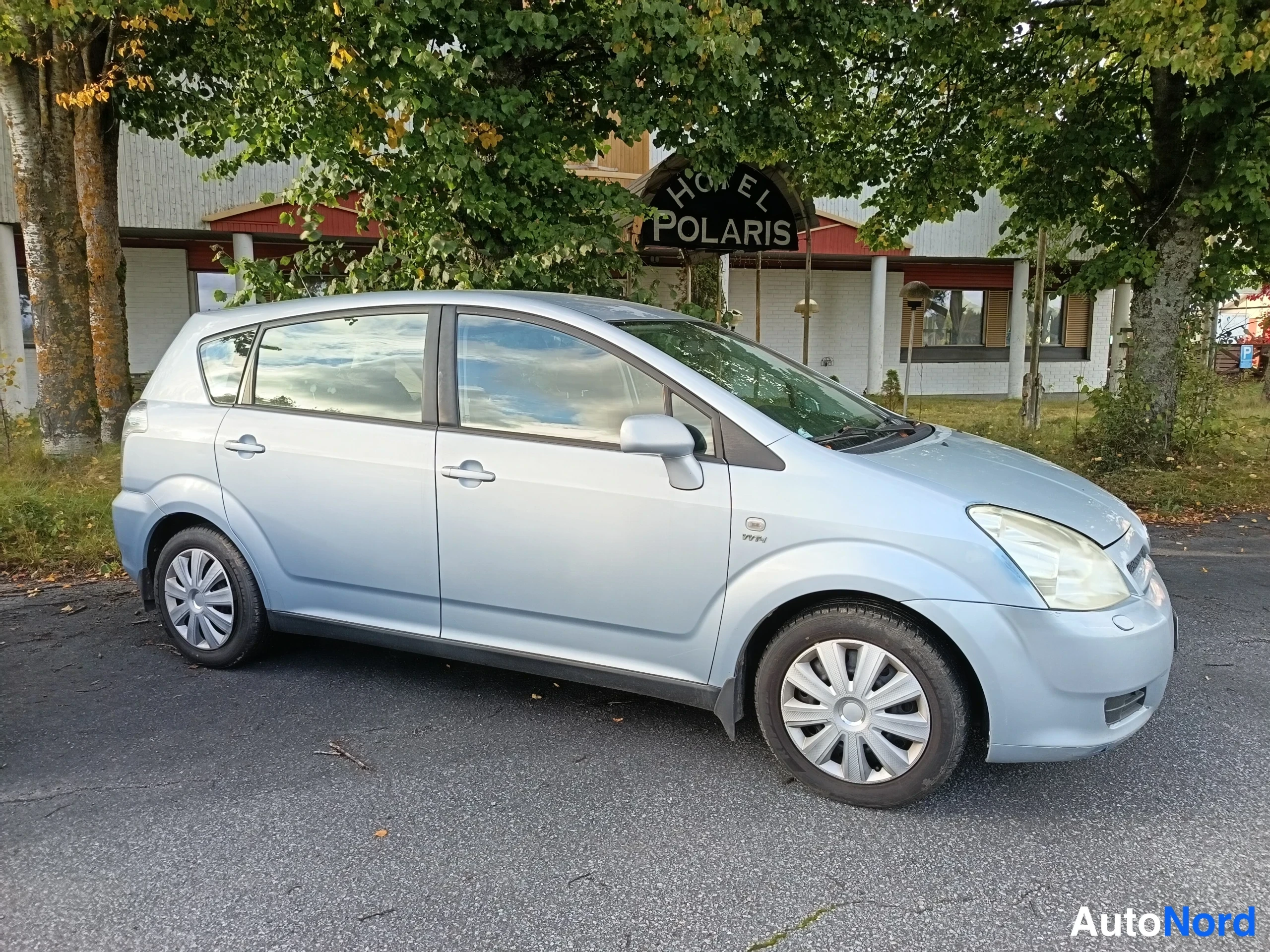 Etuviisto, nokka vasemmalle: Vaaleansininen Toyota Corolla Verso -henkilöauto sivulta etukulmasta, taustalla Hotell Polaris -rakennus ja puita.