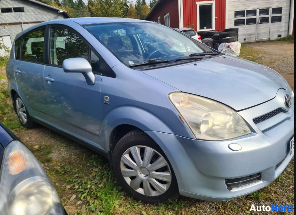 Etuviisto, nokka oikealle: Vaaleansinisen Toyota Corolla Verso -henkilöauton etukulma oikealta, edessä ovipaneelit ja konepelti, taustalla punainen rakennus ja renkaita.