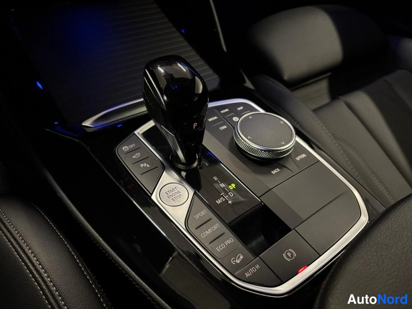 Yksityiskohta: Keskikonsolin vaihteenvalitsin ja iDrive-säädin lähikuvassa; napit Sport, Comfort, Eco Pro ja Auto H sekä Start/Stop-painike.