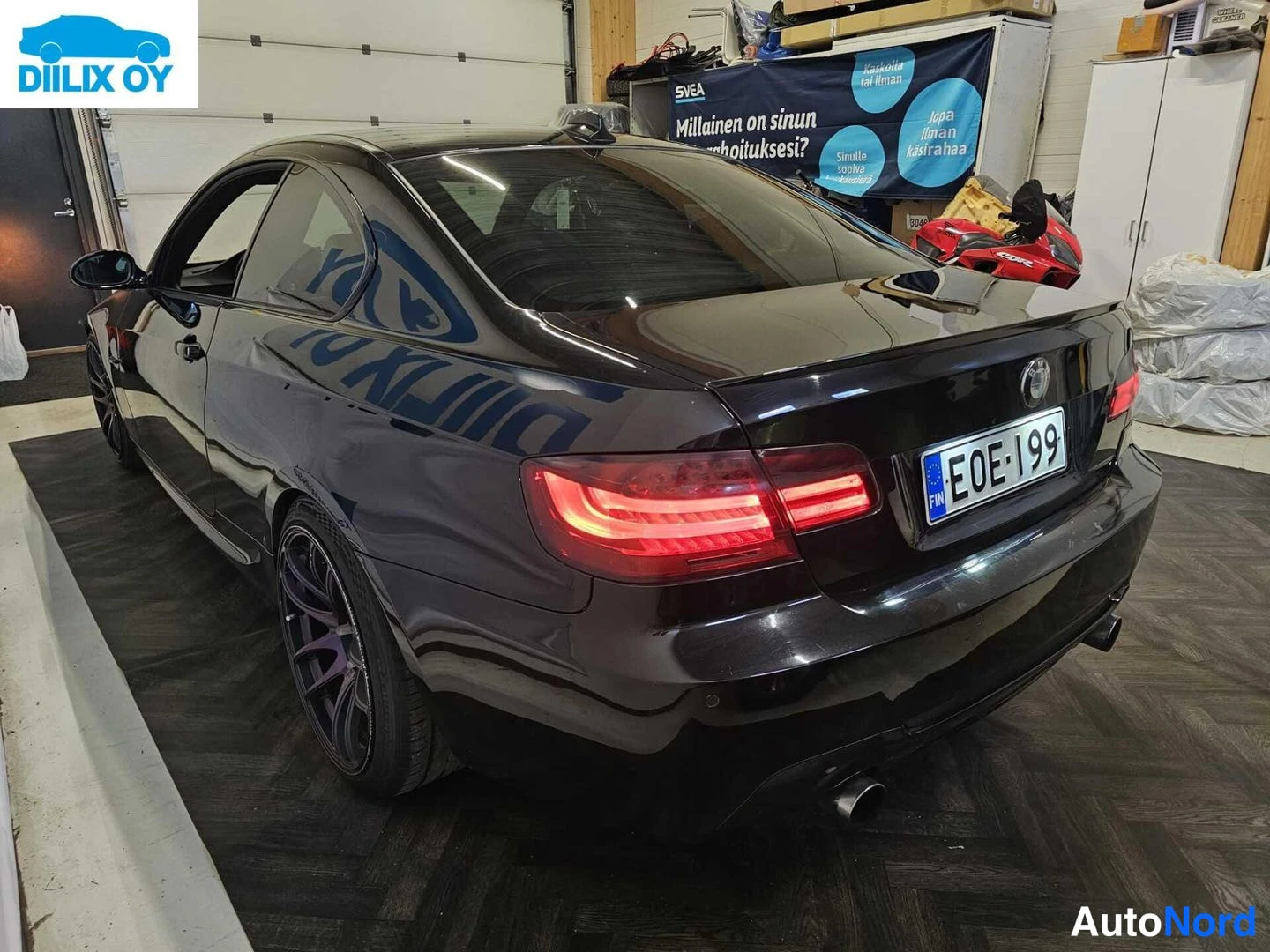 Takaviisto: Taakse ja vasemmalle suuntautuva takana näkyvä musta BMW-kupé sisätiloissa. Auton punaiset takavalot ovat päällä, rekisterikilpi on luettavissa. Seinässä on mai