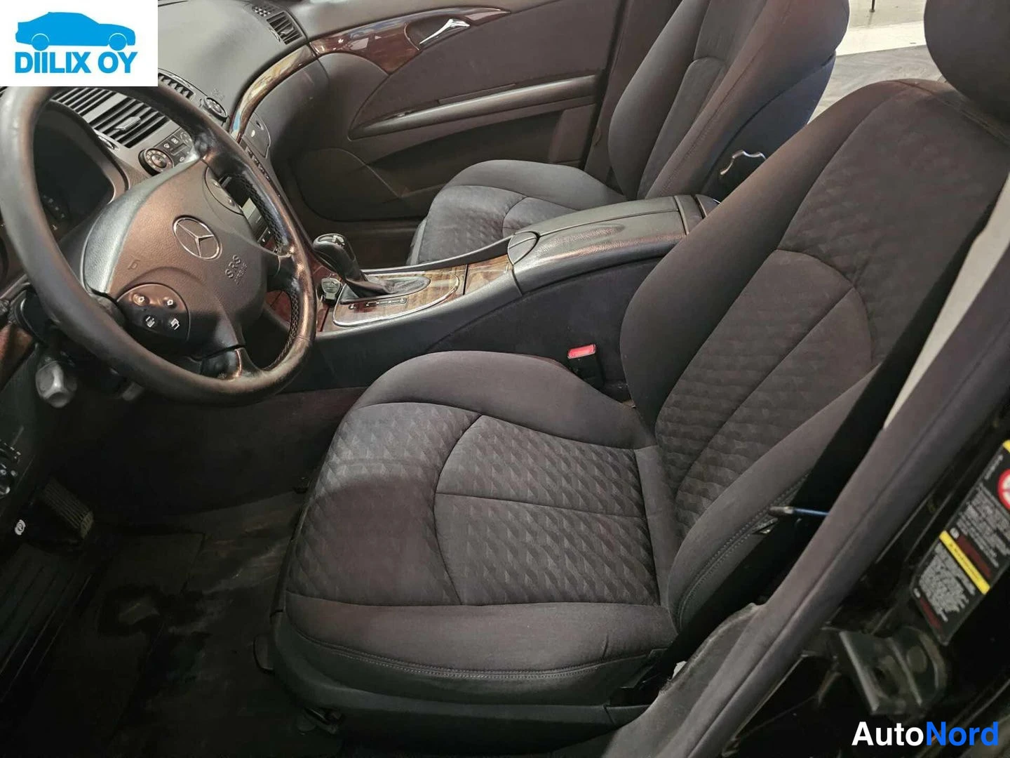 Ohjaamo: Musta Mercedes-Benz E-sarjan sisätilat. Ohjauspöydässä multifunktio-ohjauspyörä ja keskikonsolissa vaihteinvipua.