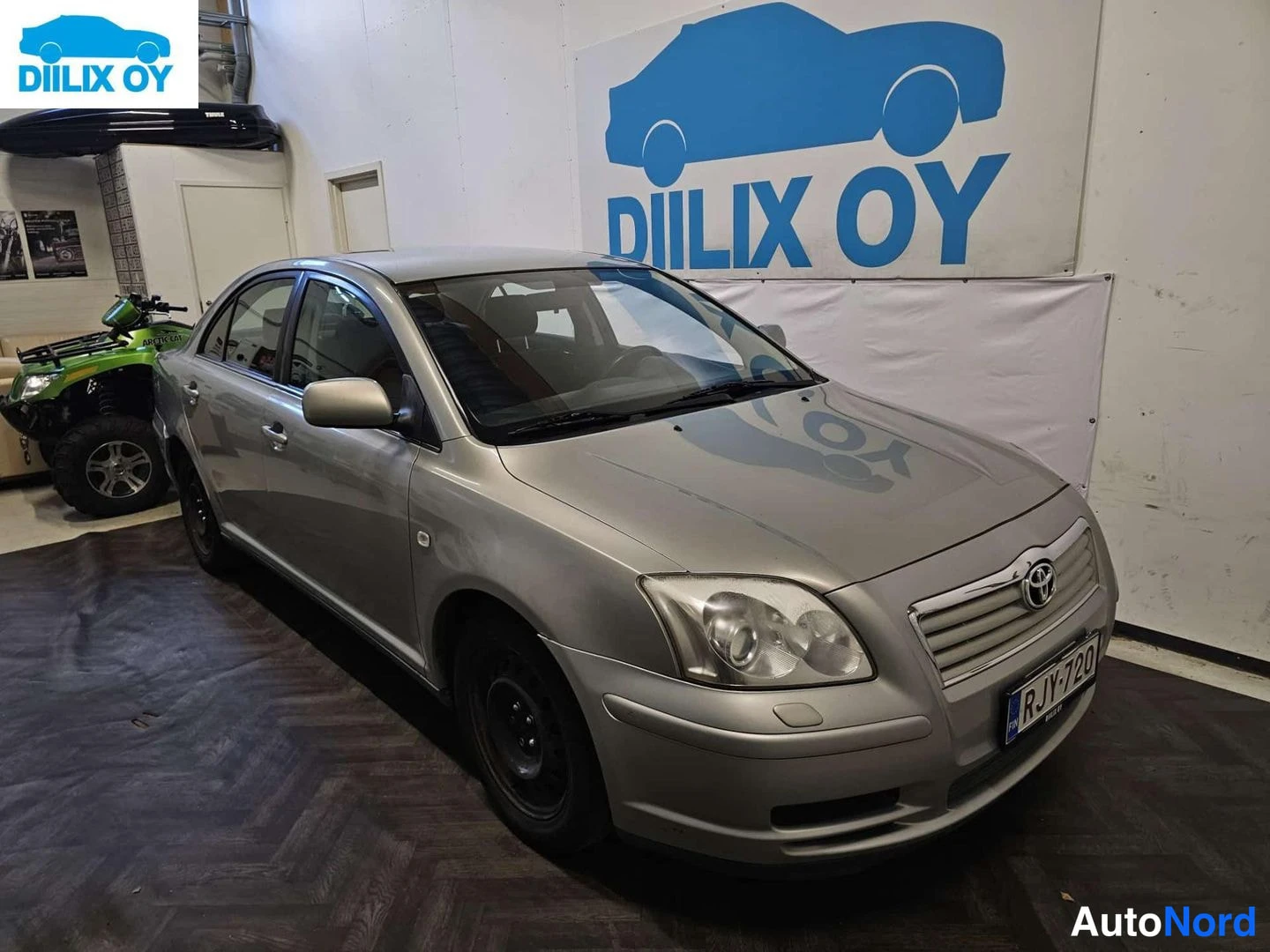 Etuviisto, nokka oikealle: Etusivun kuva.hopeanharmaa Toyota Avensis seisoo sisätiloissa; grilla, oikea etuvalo ja etupuskuri näkyvät sekä auton oikea kylki. Rekisterinumero on luk