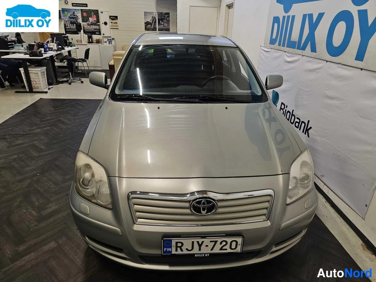 Edestä: Toyota Avensis -henkilöauto suoraan edestä kuvattuna myyntitiloissa; etusäleikkö Toyota-tunnuksella, ajovalot ja rekisterikilpi näkyvät.