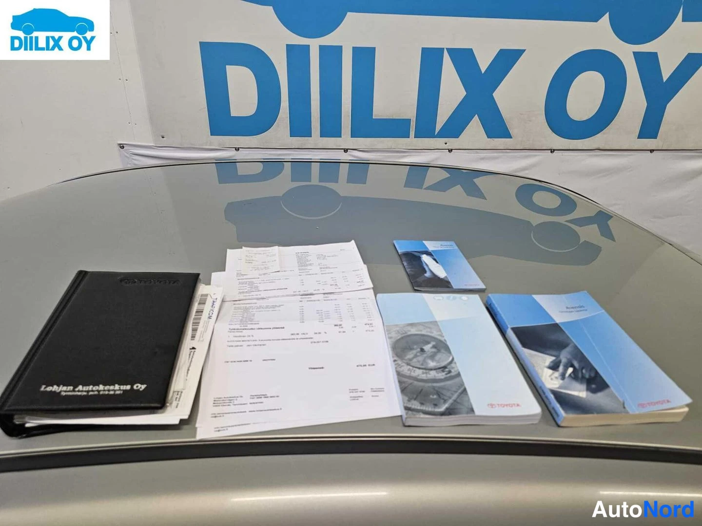 Muu: Auton harmaalla konepellillä musta kansio, paperilaskuja ja Toyota-ohjekirjoja; taustalla sininen DIILIX OY -mainoskyltti.