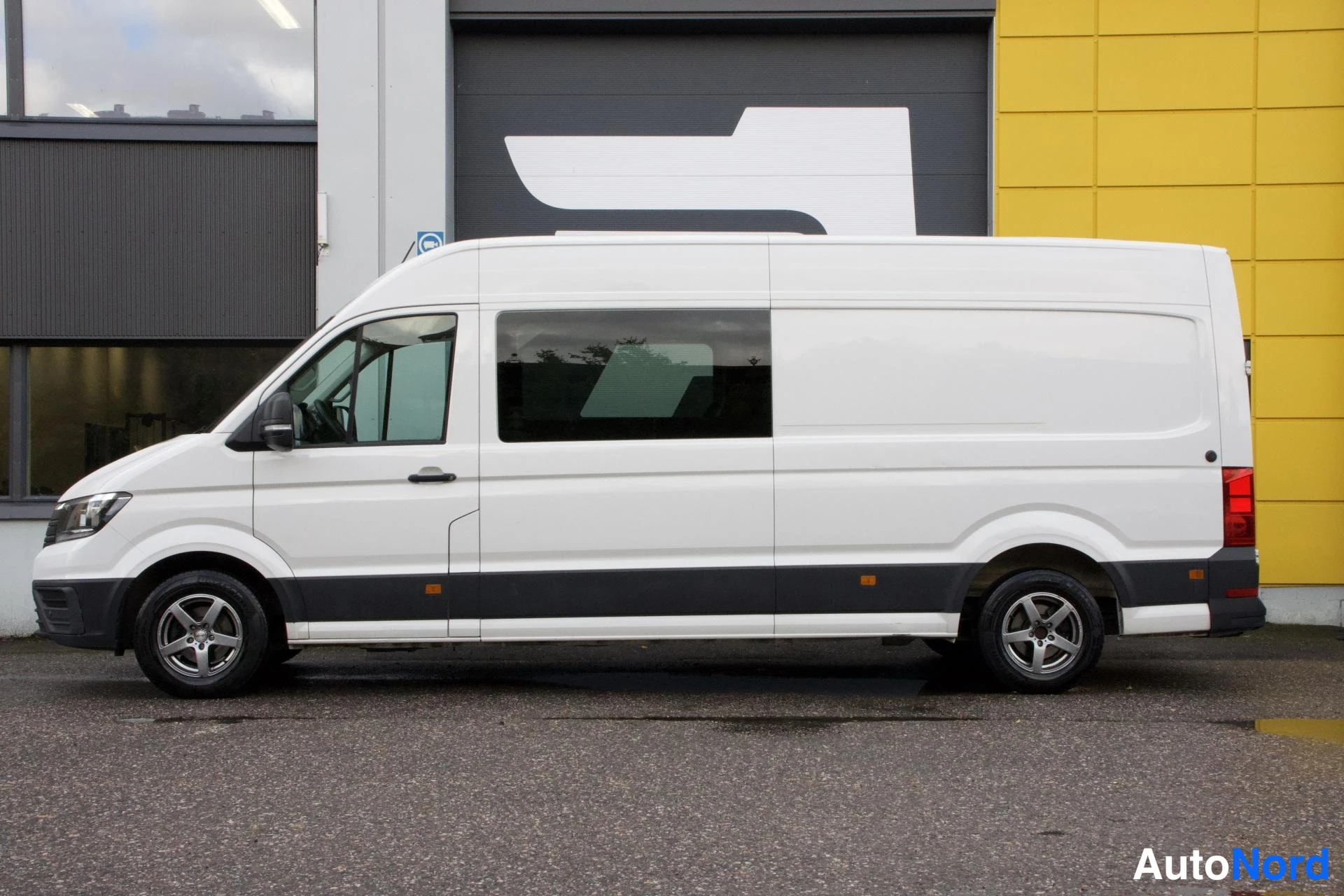 Sivulta: Valkoinen Volkswagen Crafter -pakettiauto kuvattuna vasemmalta sivulta, takapään lasiseinä ja perävaunun kiinnitys näkyvissä.