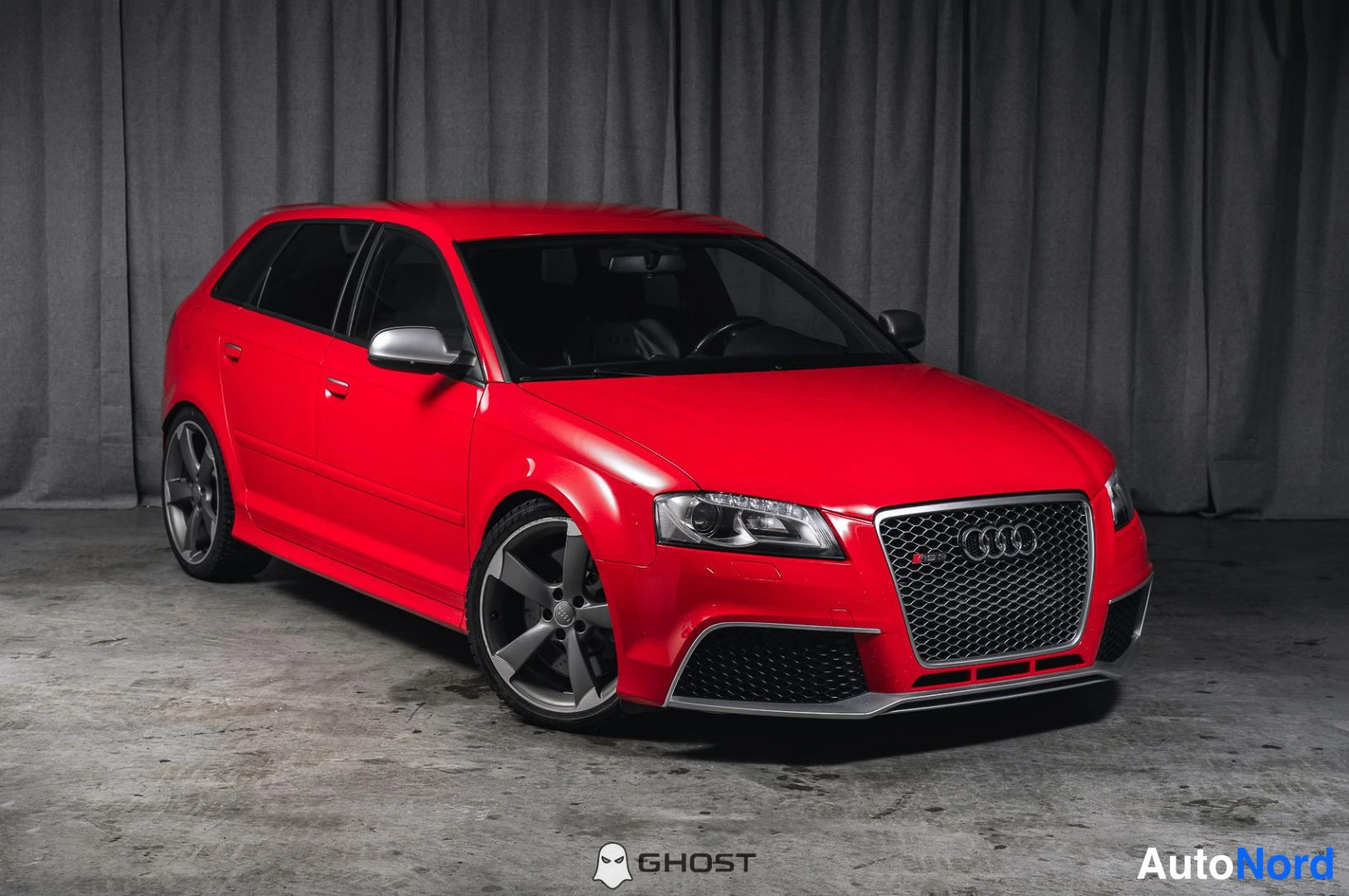 Etuviisto, nokka oikealle: Punainen Audi RS3 Sportback kolmesta neljäsosasta edestä oikeaan, harmaa verhopohja taustalla.