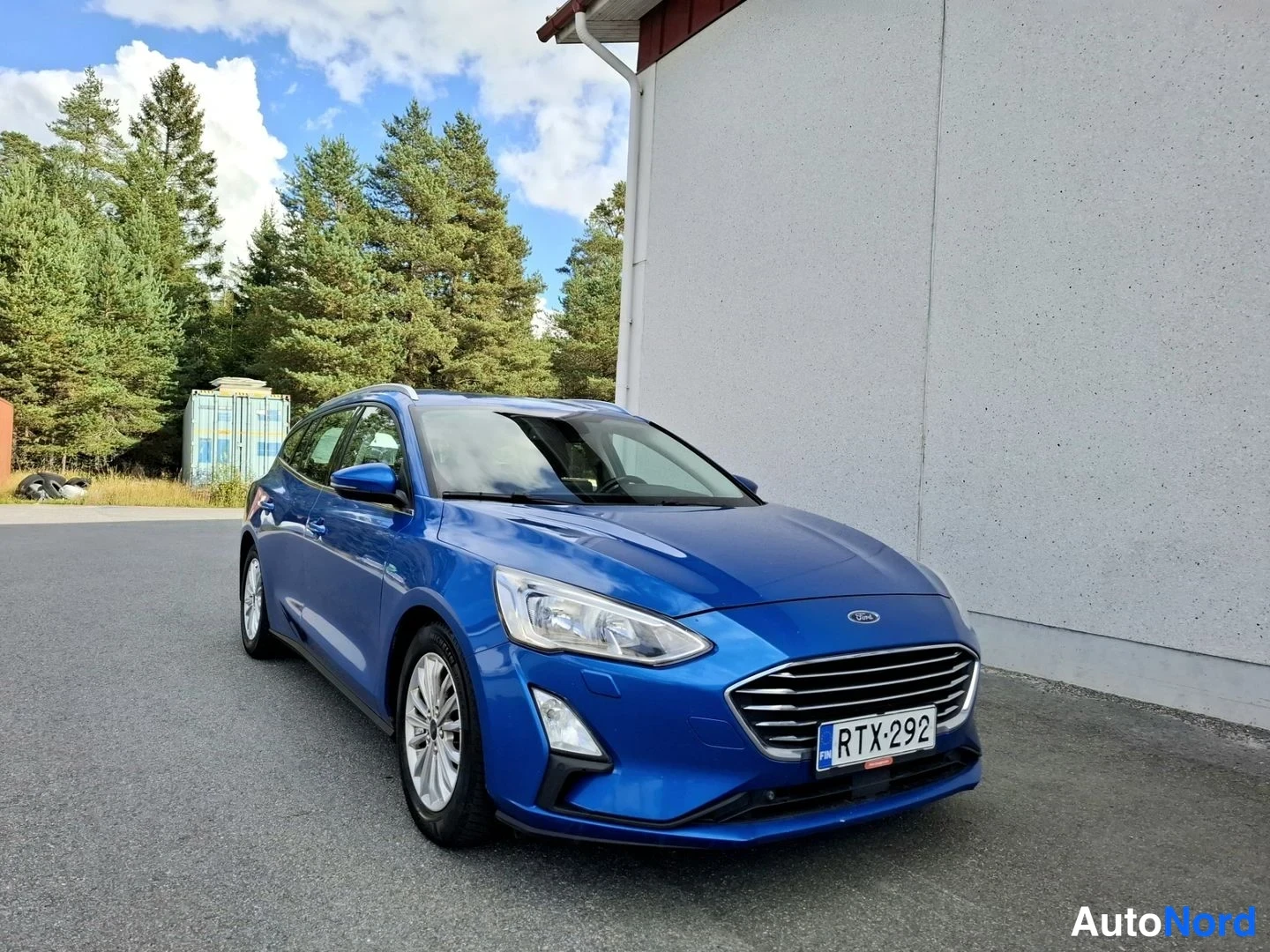 Etuviisto, nokka oikealle: Sininen Ford Focus -farmari edestä toisesta kulmasta, rekisterikilpi, valkoinen rakennus ja metsä taustalla.