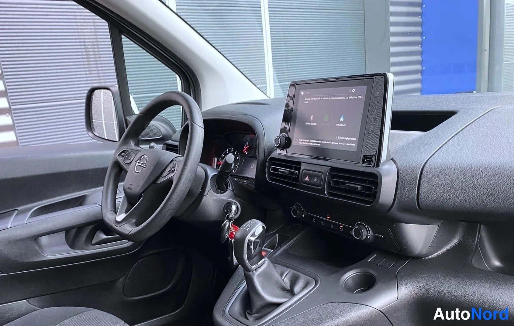 Muu: Ohjaamon näkymä kuljettajan puolelta: Opel-ohjauspyörä, kosketusnäyttö, manuaalivaihde ja avaimet kivassa.