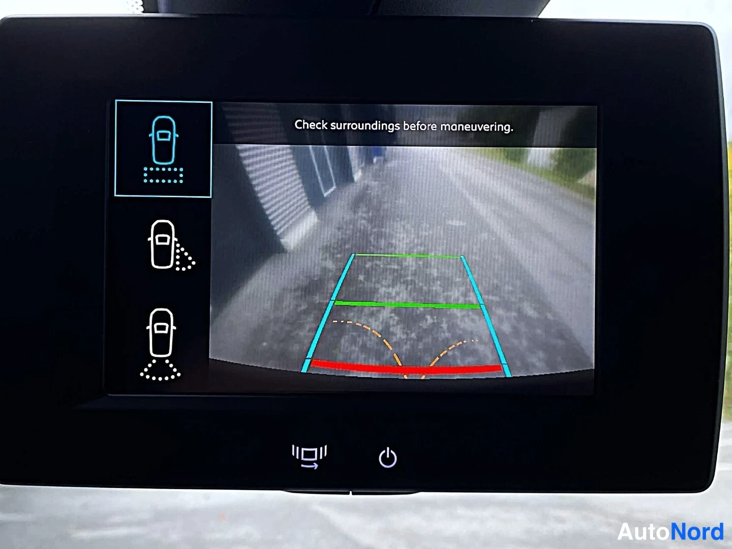 Yksityiskohta: Taaksepäin-näyttö kertoo varoituksen 'Check surroundings before maneuvering' ja näyttää kamerakuvan parkkitiloista ohjausviivoineen.